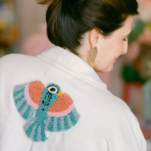 May include: A white denim jacket with a colorful embroidered bird on the back. The bird is made of yarn and has a teal blue body, pink and orange wings, and a black eye.