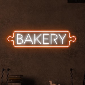 May include: A white neon sign with an orange outline that says "BAKERY" in a stylized font. The sign is mounted on a dark brown wall.