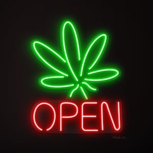 May include: A neon sign featuring a green cannabis leaf above the red word "OPEN". The sign glows against a black backdrop, creating a striking contrast. The sign is likely for a cannabis dispensary or similar establishment.