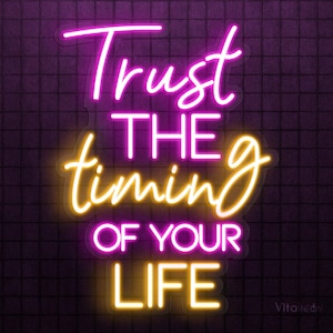 Puede incluir: Letrero de neón con la frase "Trust the timing of your life" en rosa brillante y amarillo. El letrero está montado sobre un fondo morado oscuro con un patrón de cuadrícula. Las palabras están escritas en una fuente cursiva.