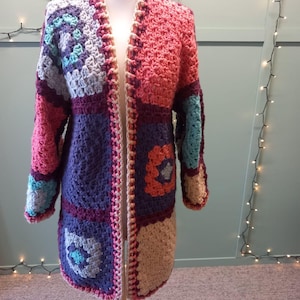 May include: A long-sleeved cardigan constructed from various crocheted granny squares in shades of coral, deep purple, light blue, and cream. The garment features an open front design with a cream-coloured crocheted border along the edges.