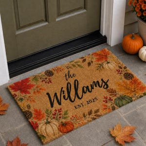 May include: A rectangular tan coir doormat featuring a printed border of colorful autumn leaves, pumpkins, and pinecones. The center displays the black script text "the Willams" above the printed text "EST. 2025" in a simple sans-serif font.