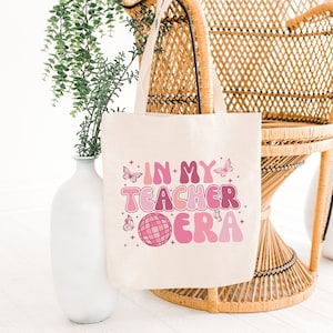 Könnte beinhalten: Eine beigefarbene Tragetasche mit dem Text "IN MY TEACHER ERA" in rosa und lila Retro-Schrift. Die Tasche hat Schmetterlings- und Discokugel-Grafiken. Die Tragetasche hat zwei Griffe und wird auf einem Korbstuhl präsentiert.