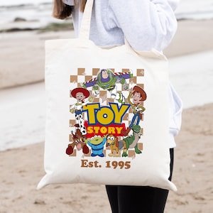 May include: White canvas tote bag with a brown and white checkered background. The bag features a graphic of the Toy Story characters, including Woody, Buzz Lightyear, and Rex. The text "Toy Story Est. 1995" is printed on the bag.