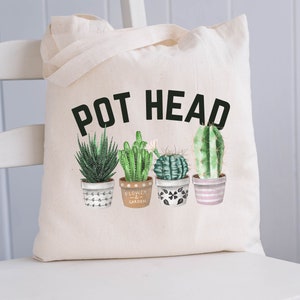 May include: A white canvas tote bag with the text "POT HEAD" in black letters. The bag features a graphic of four potted succulents in various shades of green.