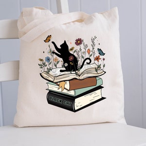 Könnte beinhalten: Eine weiße Einkaufstasche mit einer schwarzen Katze, die auf einem Stapel Bücher sitzt. Die Katze streckt die Pfote nach einem Schmetterling aus. Die Bücher sind von Blumen umgeben und das oberste Buch trägt den Schriftzug "Black Cat".