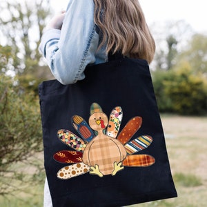 May include: A black tote bag featuring a patchwork turkey design. The turkey is made from various fabrics, including plaid, floral and polka dot patterns. The turkey is brown, orange and yellow.