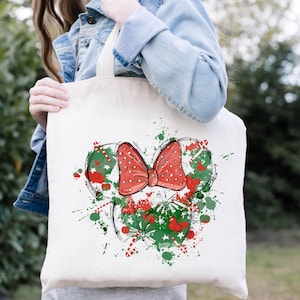 Puede incluir: Una bolsa de lona blanca con un lazo rojo con lunares blancos y un dise&ntilde;o navide&ntilde;o verde y rojo. El dise&ntilde;o es una silueta estilizada de la cabeza de Mickey Mouse.