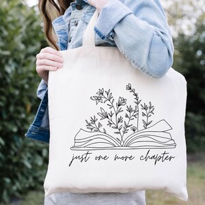 K&ouml;nnte beinhalten: Eine wei&szlig;e Einkaufstasche mit einer schwarzen Linienzeichnung eines offenen Buches, aus dem Blumen wachsen. Der Text "just one more chapter" ist unter dem Buch gedruckt.