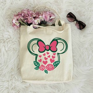 Puede incluir: Una bolsa de lona blanca con una silueta verde y rosa de una cabeza de rat&oacute;n con un lazo y corazones. La cabeza del rat&oacute;n est&aacute; rodeada de hojas verdes y rosas rosas.