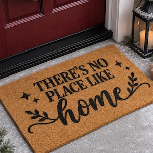 May include: A rectangular tan coir doormat featuring the black text "THERE'S NO PLACE LIKE home" in a mix of serif and script fonts. The design includes small four-pointed star accents and decorative leafy vine flourishes on the sides.