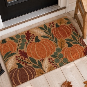 Peut inclure: Un paillasson rectangulaire en fibre de coco avec un motif imprimé de citrouilles orange et marron, de feuilles vert foncé et de petites baies rouges. Le design est disposé sur la surface en fibre naturelle beige, créant un motif automnal.