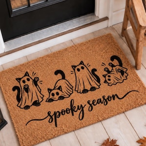 Peut inclure: Un paillasson rectangulaire en fibre de coco beige avec quatre illustrations noires de chats déguisés en fantômes. Le texte "spooky season" est imprimé en écriture cursive noire sous les chats. Le tapis a une texture fibreuse naturelle.