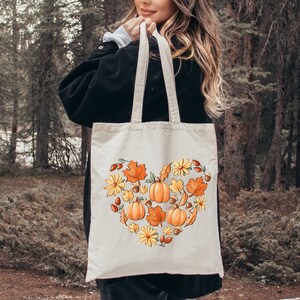 May include: A white canvas tote bag with a heart-shaped design featuring orange pumpkins, yellow flowers, brown acorns, and red berries. The design is surrounded by green leaves.