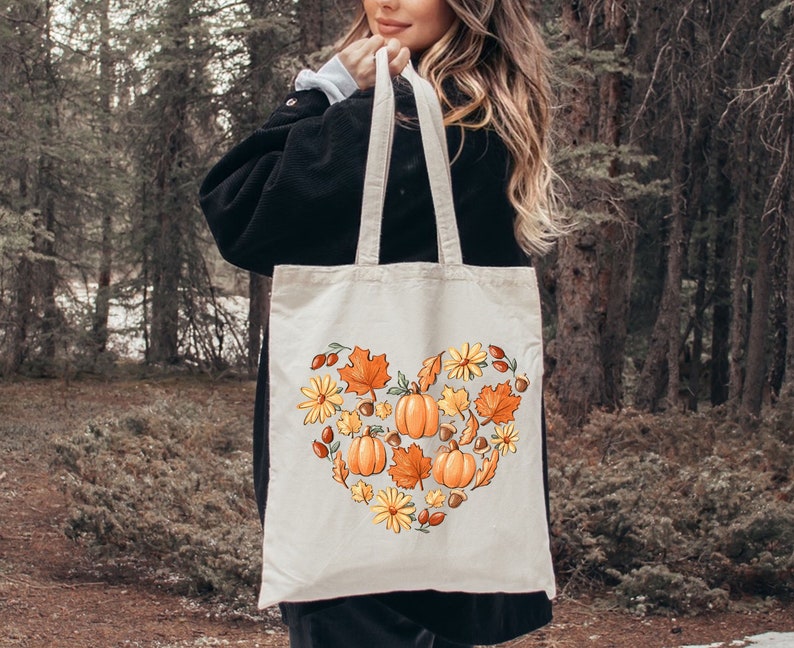 May include: A white canvas tote bag with a heart-shaped design featuring orange pumpkins, yellow flowers, brown acorns, and red berries. The design is surrounded by green leaves.