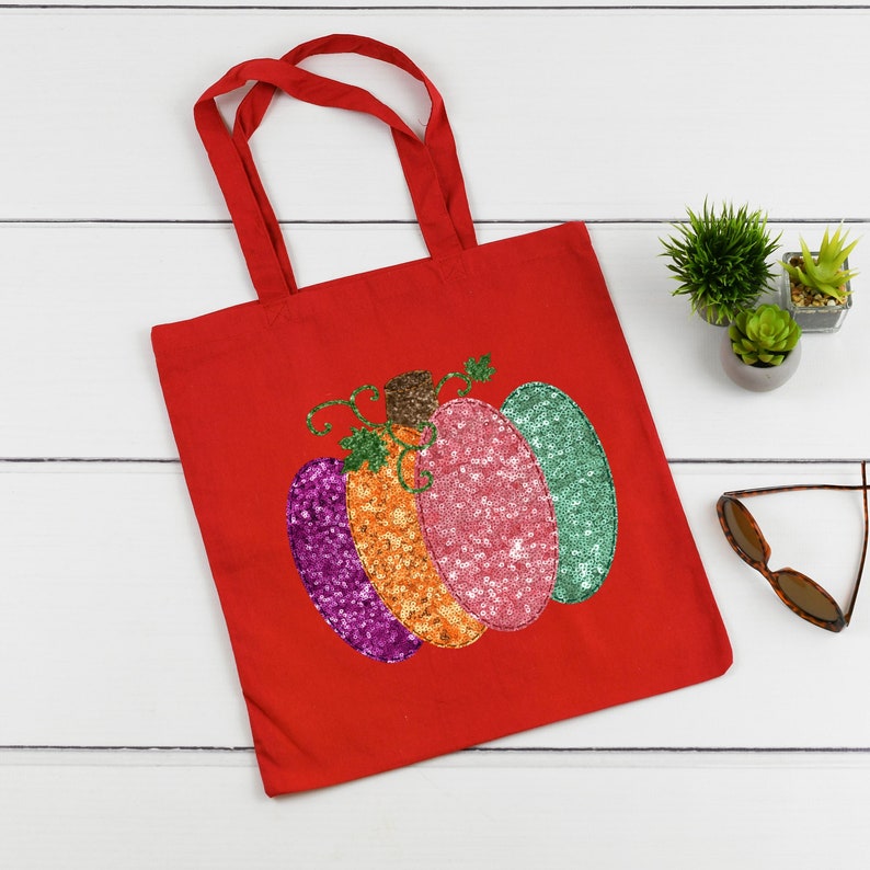 May include: Red canvas tote bag with a sequined pumpkin design. The pumpkin is multi-colored with pink, orange, green, and purple sequins.