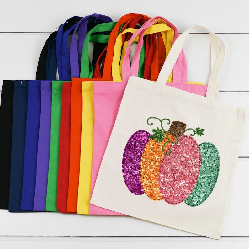 May include: A set of 10 colorful tote bags with a single bag featuring a sparkly pumpkin design. The bags are made of canvas and have a variety of colors including red, yellow, green, blue, and purple.