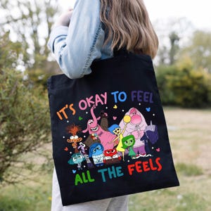 K&ouml;nnte beinhalten: Schwarze Einkaufstasche mit einer bunten Grafik von Figuren aus dem Film Inside Out. Der Text "Es ist okay, alle Gef&uuml;hle zu f&uuml;hlen" ist auf die Tasche gedruckt.