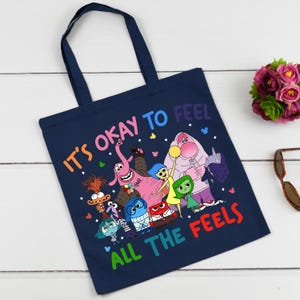 K&ouml;nnte beinhalten: Eine marineblaue Tragetasche mit einer bunten Grafik von Figuren aus dem Film Inside Out. Der Text "It's okay to feel all the feels" ist auf die Tasche gedruckt.