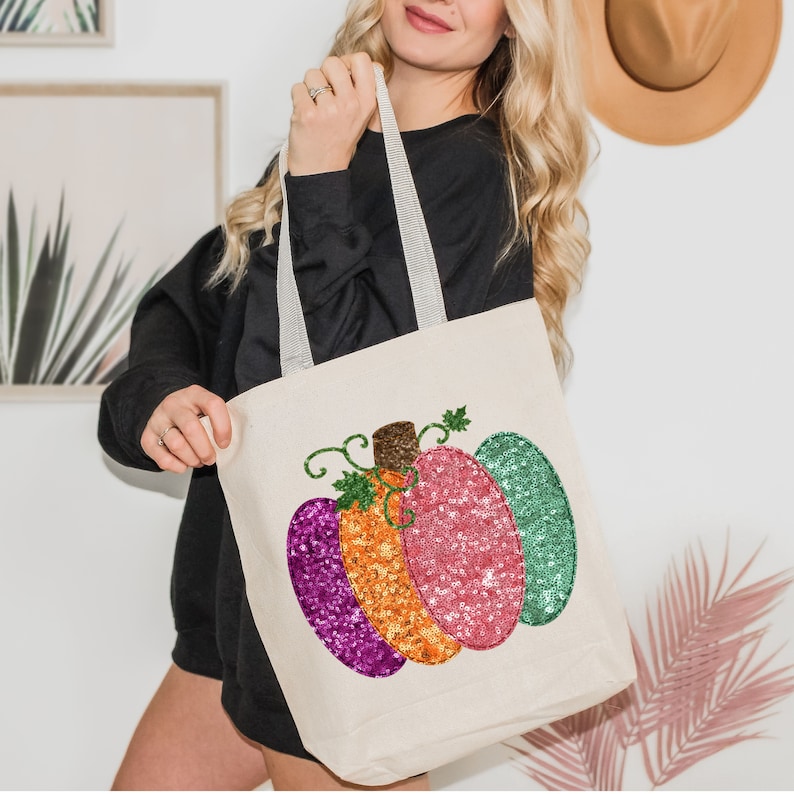 May include: A white canvas tote bag with a colorful sequined pumpkin design. The pumpkin is made up of three sections: a purple section, an orange section, and a green section. The pumpkin has a green stem and leaves.