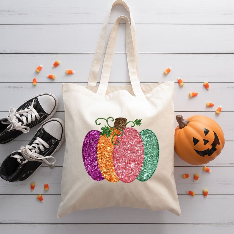 May include: A white canvas tote bag with a glittery pumpkin design in shades of purple, orange, pink, and green. The pumpkin has a brown stem and green leaves.