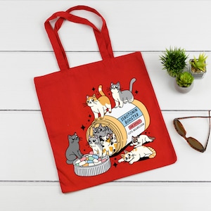 May include: Red tote bag with a colorful illustration of cats inside a pill bottle labeled "Serotonin Booster". The side effect is listed as "Cute Aggression".