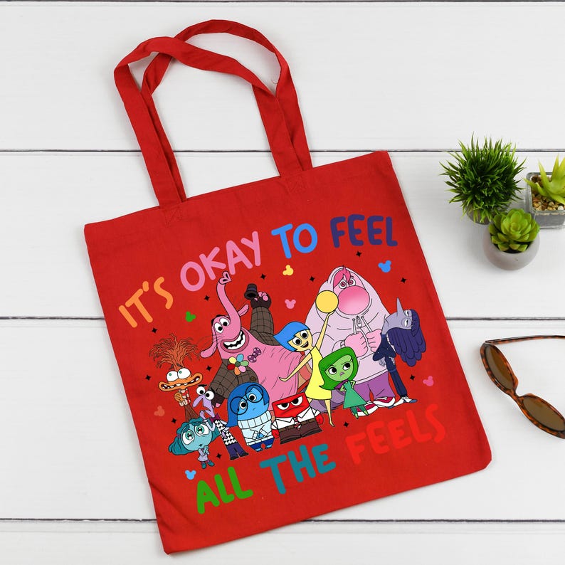 K&ouml;nnte beinhalten: Rote Stofftasche mit einem bunten Grafikdruck von Figuren aus dem Film Inside Out. Der Text auf der Tasche lautet "Es ist okay, alle Gef&uuml;hle zu f&uuml;hlen."