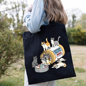 May include: Black tote bag with a white design of a pill bottle labeled "Serotonin Booster" with a side effect of "Cute Aggression".  The pill bottle is overflowing with colorful pills and several cartoon cats are spilling out of the bottle.