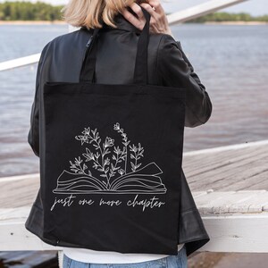 K&ouml;nnte beinhalten: Schwarze Einkaufstasche mit einer wei&szlig;en Umrandung eines offenen Buches, aus dem Blumen wachsen. Der Text "just one more chapter" ist unter dem Buch gedruckt.