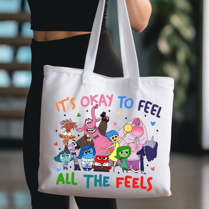 K&ouml;nnte beinhalten: Wei&szlig;e Canvas-Tragetasche mit dem Text "Es ist okay, alle Gef&uuml;hle zu f&uuml;hlen" in Regenbogenfarben. Die Tasche zeigt eine bunte Illustration von Figuren aus dem Film Inside Out.