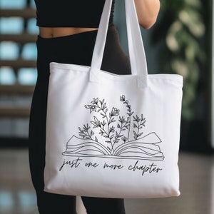K&ouml;nnte beinhalten: Wei&szlig;e Canvas-Tragetasche mit schwarzer Linienzeichnung eines offenen Buches, aus dem Blumen wachsen. Der Text "just one more chapter" ist unter dem Buch gedruckt.