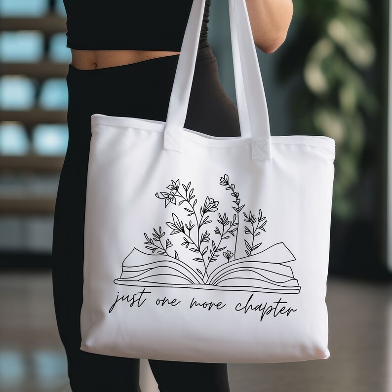 K&ouml;nnte beinhalten: Wei&szlig;e Canvas-Tragetasche mit schwarzer Linienzeichnung eines offenen Buches, aus dem Blumen wachsen. Der Text "just one more chapter" ist unter dem Buch gedruckt.