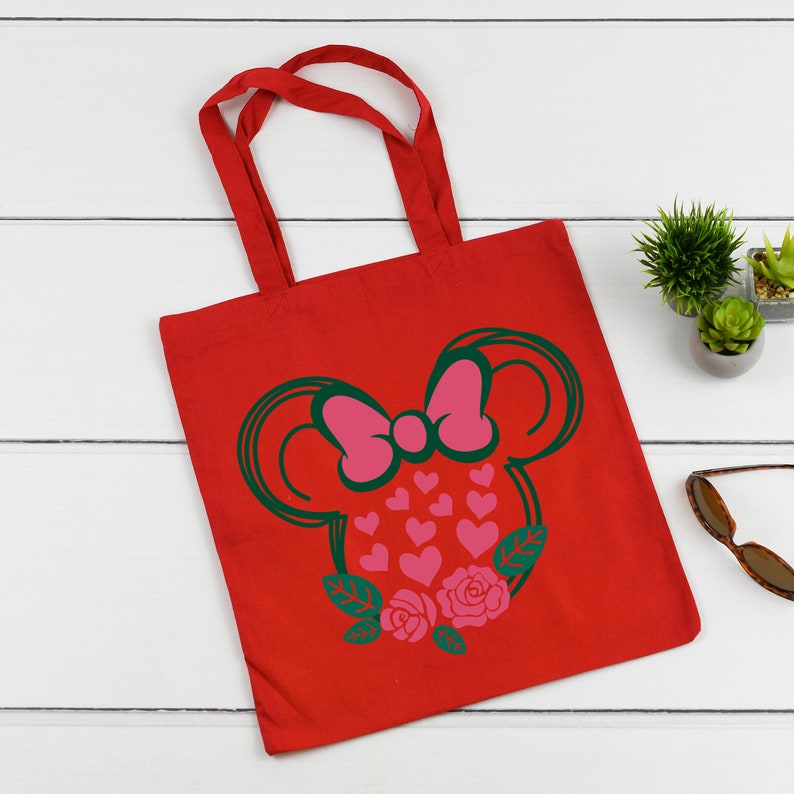 Puede incluir: Bolsa de lona roja con una silueta verde y rosa de una cabeza de rat&oacute;n con un lazo y corazones. La cabeza del rat&oacute;n est&aacute; rodeada de hojas verdes y rosas rosas.