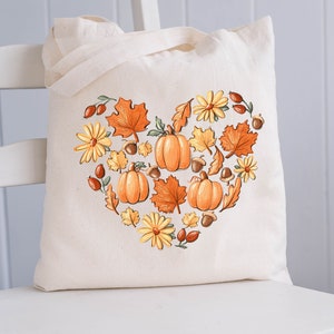 May include: A white canvas tote bag with a heart-shaped design featuring pumpkins, leaves, and flowers in shades of orange, yellow, and brown. The design is reminiscent of autumn.