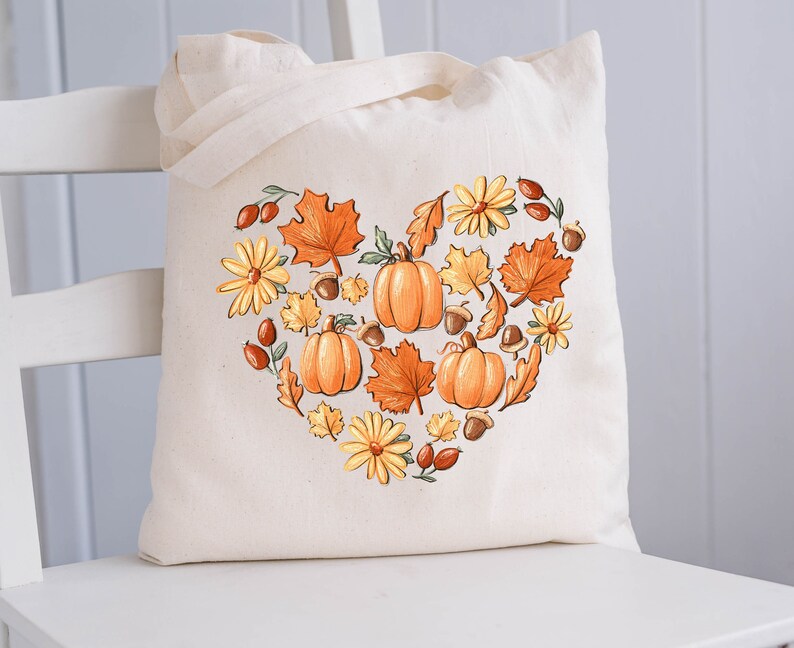 May include: A white canvas tote bag with a heart-shaped design featuring pumpkins, leaves, and flowers in shades of orange, yellow, and brown. The design is reminiscent of autumn.