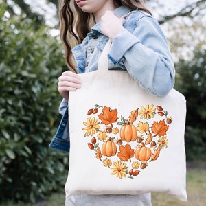 May include: A white tote bag with a heart-shaped design featuring fall-themed elements like pumpkins, leaves, acorns, and flowers. The design is in shades of orange, yellow, and brown.
