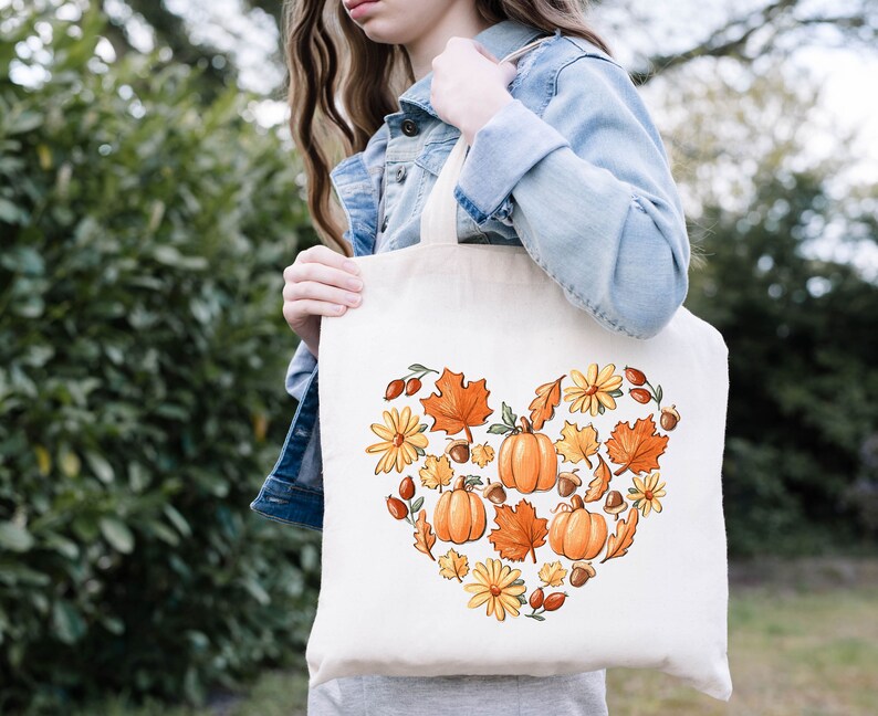 May include: A white tote bag with a heart-shaped design featuring fall-themed elements like pumpkins, leaves, acorns, and flowers. The design is in shades of orange, yellow, and brown.