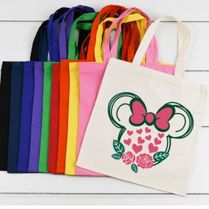 Puede incluir: Un conjunto de 10 bolsas de tela en blanco en varios colores, incluyendo azul marino, morado, verde, rojo, amarillo y rosa. La &uacute;ltima bolsa es blanca con un contorno verde de una cabeza de rat&oacute;n de dibujos animados con un lazo rosa y corazones.