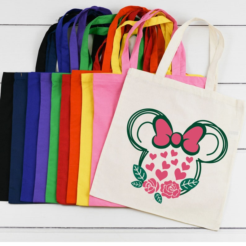 Puede incluir: Un conjunto de 10 bolsas de tela en blanco en varios colores, incluyendo azul marino, morado, verde, rojo, amarillo y rosa. La &uacute;ltima bolsa es blanca con un contorno verde de una cabeza de rat&oacute;n de dibujos animados con un lazo rosa y corazones.