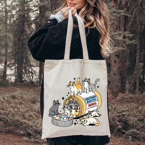 May include: A white tote bag with a cartoon illustration of a pill bottle filled with cats. The text on the bottle reads "Serotonin Booster" and "Side Effect: Cute Aggression".