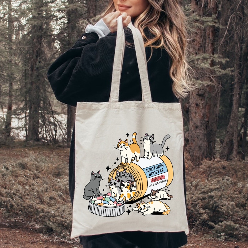 May include: A white tote bag with a cartoon illustration of a pill bottle filled with cats. The text on the bottle reads "Serotonin Booster" and "Side Effect: Cute Aggression".