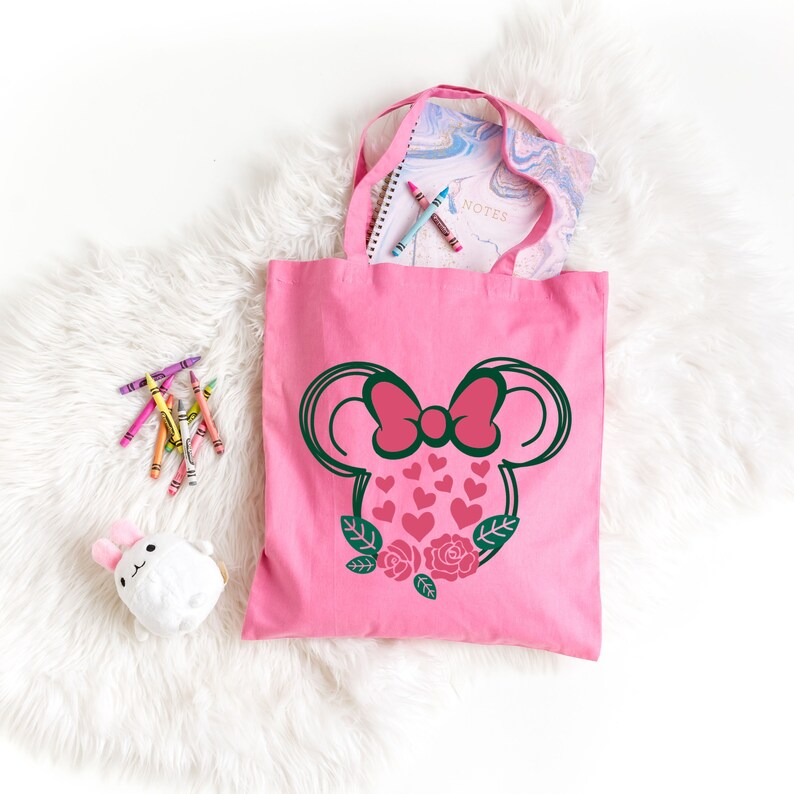 Puede incluir: Bolsa de lona rosa con una silueta rosa y verde de una cabeza de rat&oacute;n con un lazo y corazones. La cabeza del rat&oacute;n est&aacute; rodeada de hojas verdes y rosas rosas.
