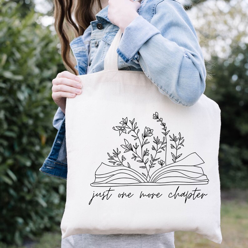 K&ouml;nnte beinhalten: Eine wei&szlig;e Einkaufstasche mit einer schwarzen Linienzeichnung eines offenen Buches, aus dem Blumen wachsen. Der Text "just one more chapter" ist unter dem Buch gedruckt.