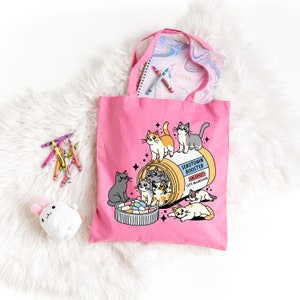 May include: Pink tote bag with a cartoon design of a pill bottle filled with cats. The pill bottle says "Serotonin Booster" and "Side Effect: Cute Aggression".