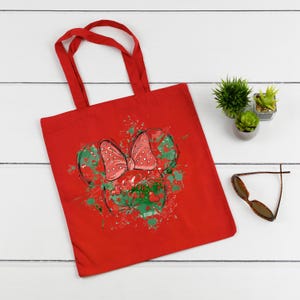 Puede incluir: Bolsa de tela roja con un dise&ntilde;o de Minnie Mouse verde y rojo. El dise&ntilde;o presenta una cabeza de Minnie Mouse verde y roja con un lazo rojo con lunares blancos. El fondo es un lienzo rojo con salpicaduras de pintura verde y roja.