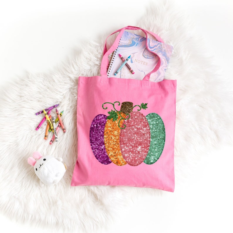 May include: Pink canvas tote bag with a sequined pumpkin design. The pumpkin is multi-colored with purple, orange, and green sequins. The bag has two handles and is perfect for carrying books, supplies, or other items.