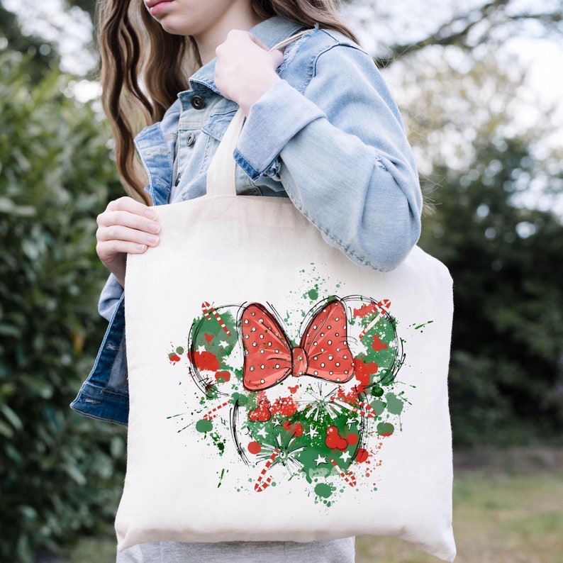Puede incluir: Una bolsa de lona blanca con un lazo rojo con lunares blancos y un dise&ntilde;o navide&ntilde;o verde y rojo. El dise&ntilde;o es una silueta estilizada de la cabeza de Mickey Mouse.