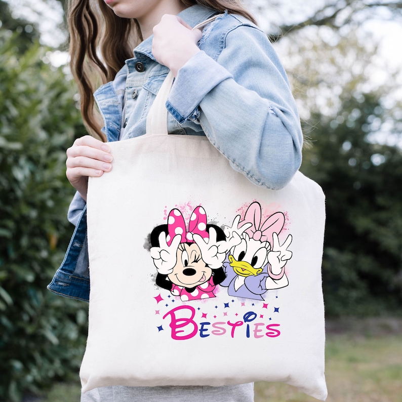 May include: A white canvas tote bag with a pink and purple design featuring Minnie Mouse and Daisy Duck. The design includes the word "Besties" in pink and purple letters.