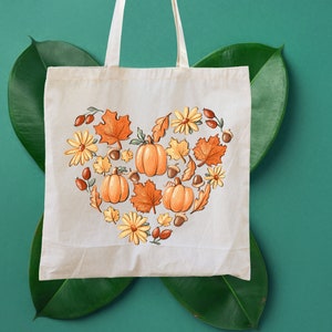 May include: A white tote bag with a heart-shaped design featuring pumpkins, leaves, and flowers in shades of orange, yellow, and brown. The design is centered on the bag and is surrounded by a white border.