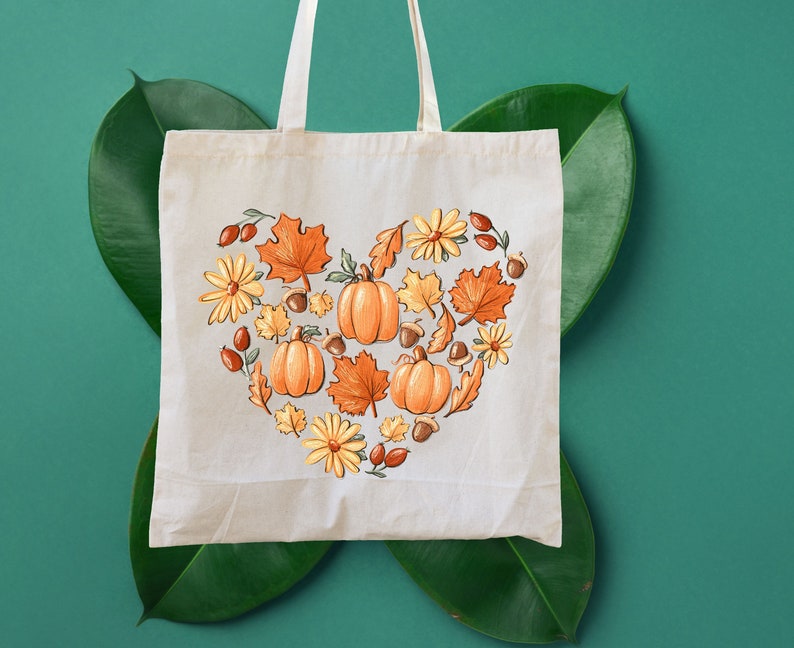 May include: A white tote bag with a heart-shaped design featuring pumpkins, leaves, and flowers in shades of orange, yellow, and brown. The design is centered on the bag and is surrounded by a white border.
